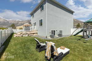 Expansive backyard featuring a manicured lawn, white privacy fencing, a raised deck with white railings, a rock retaining wall, and a mountain backdrop