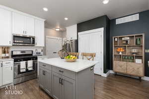 Modern kitchen featuring a central island with a quartz countertop, wood-finish flooring, white upper cabinetry, grey lower cabinetry, and stainless steel appliances including a gas range and microwave