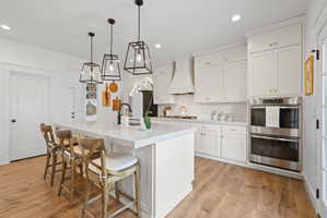 Bright kitchen featuring a substantial island with seating, pendant lighting, white shaker cabinetry, stainless steel double ovens, and wood-finish flooring