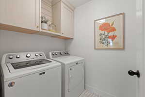 Laundry area featuring two white appliances, light-toned upper cabinetry, open shelving with shiplap backing, and white hexagonal floor tiles