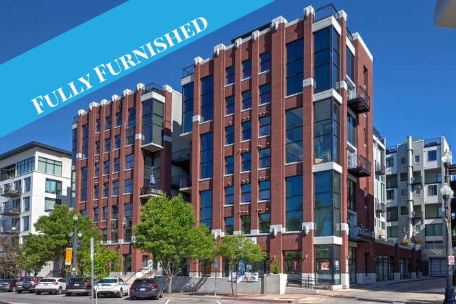 Modern multi-story building featuring extensive brickwork, large pane windows, multiple balconies with metal railings, and ground-level retail spaces