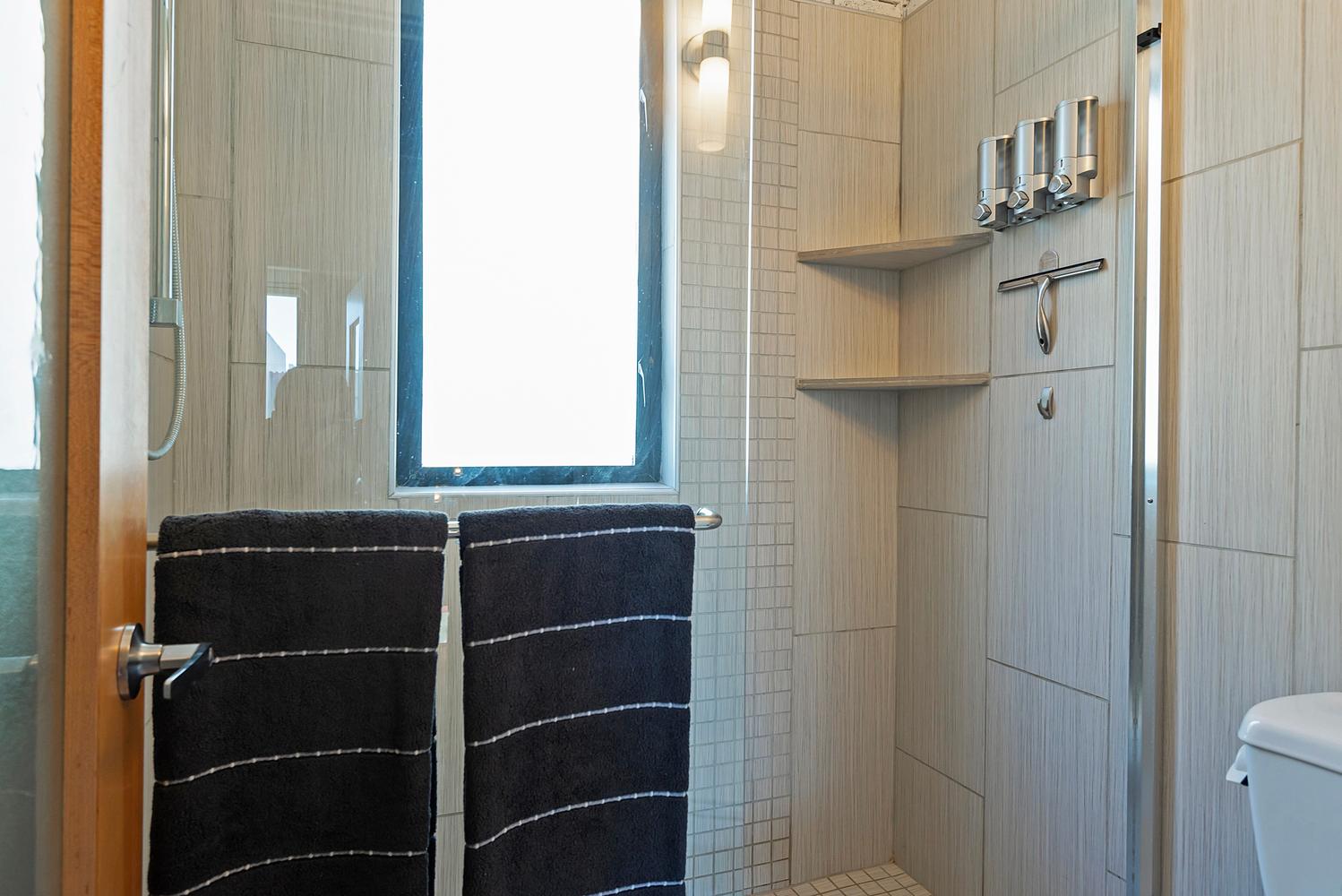 Shower enclosure featuring light-toned tile walls, built-in corner shelving, a recessed window, a wall-mounted fixture, and a glass partition