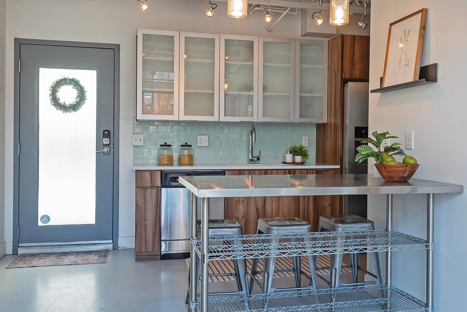 Modern kitchen featuring a stainless steel countertop island, wood-finish lower cabinetry, frosted glass upper cabinets, a turquoise subway tile backsplash, and stainless steel appliances