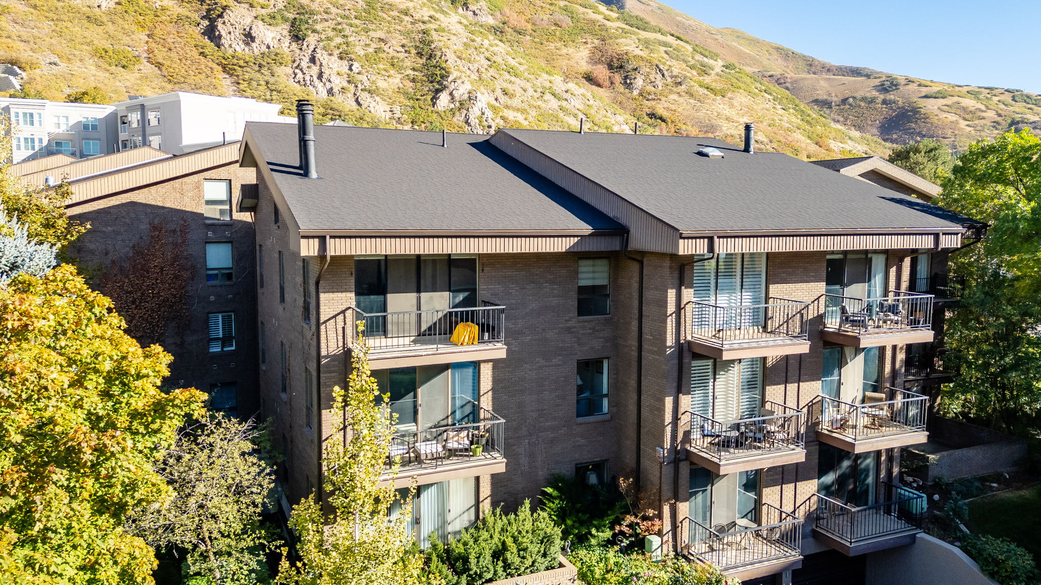 Multi-story building featuring brick and vertical siding facades, dark asphalt shingle roofing, multiple private balconies with metal railings, and a mountainous backdrop