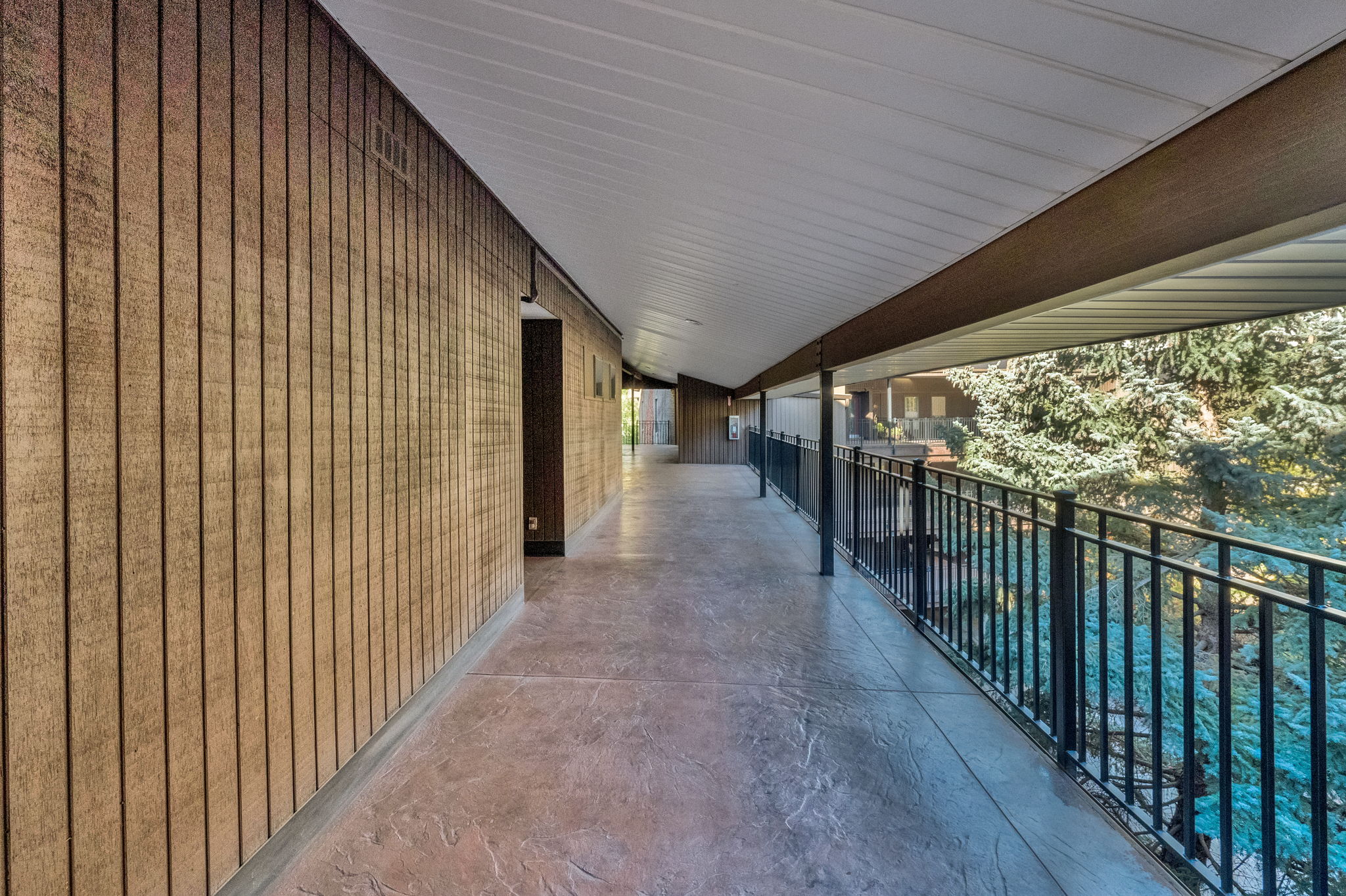 Covered outdoor walkway featuring a textured concrete surface, slatted wood-finish wall paneling, and a black metal railing