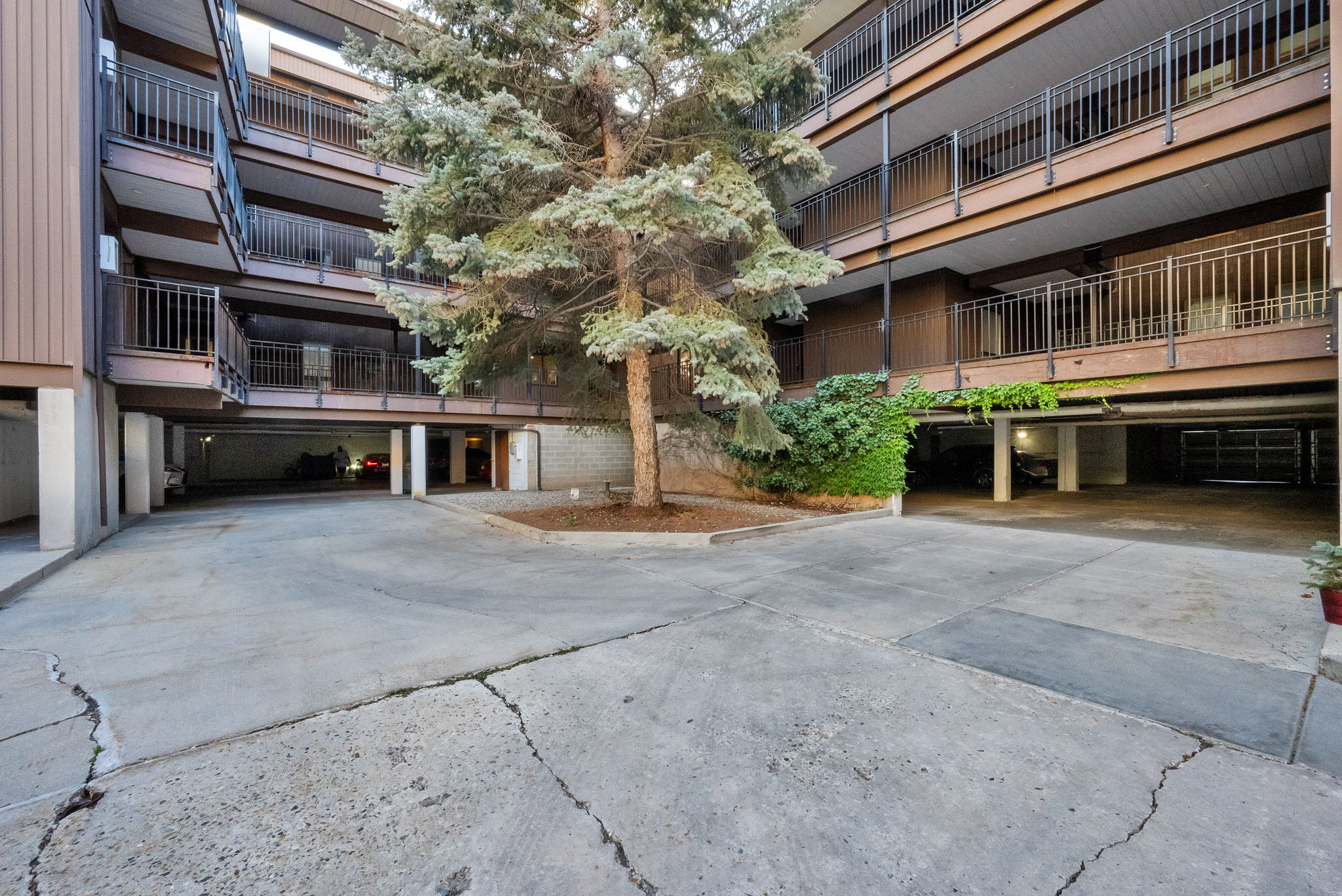 Multi-level apartment building featuring private balconies with black metal railings, a central parking area, mature landscaping, and an integrated covered parking structure
