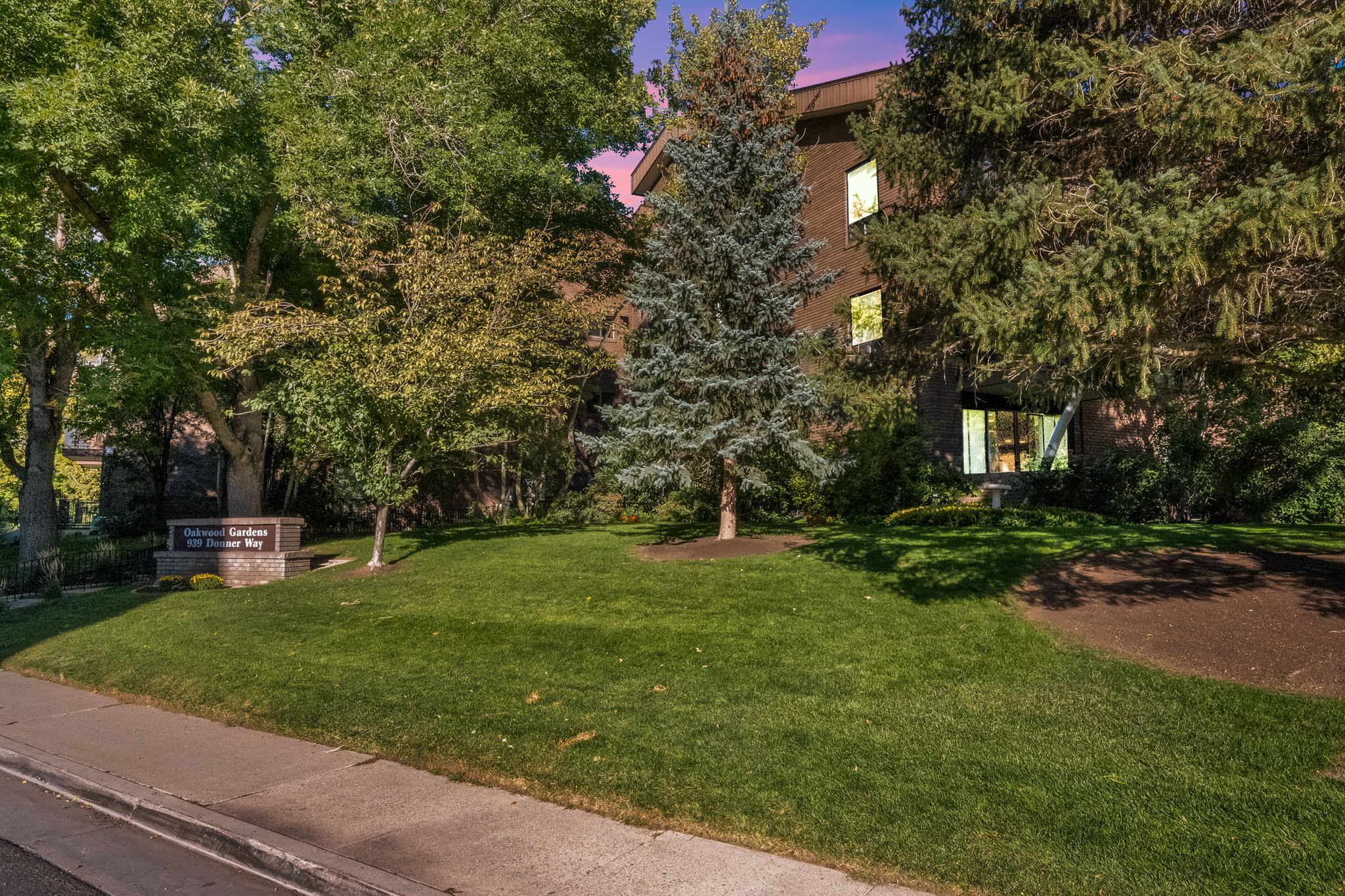 Manicured green lawn with mature trees and shrubbery, featuring a stone-clad monument sign, a paved pedestrian path, and a multi-story building facade with large windows