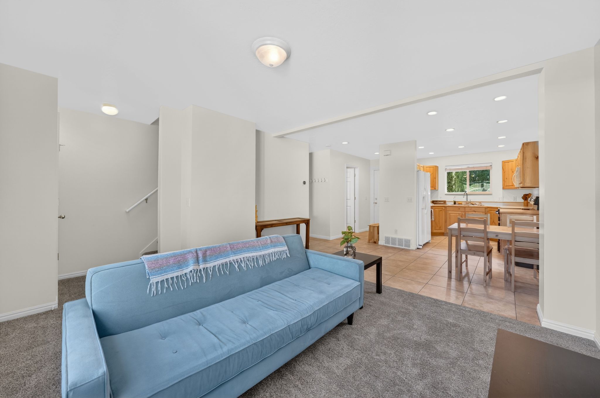 Living space featuring light gray carpeting, neutral wall tones, recessed lighting in background, and a prominent staircase with white handrail