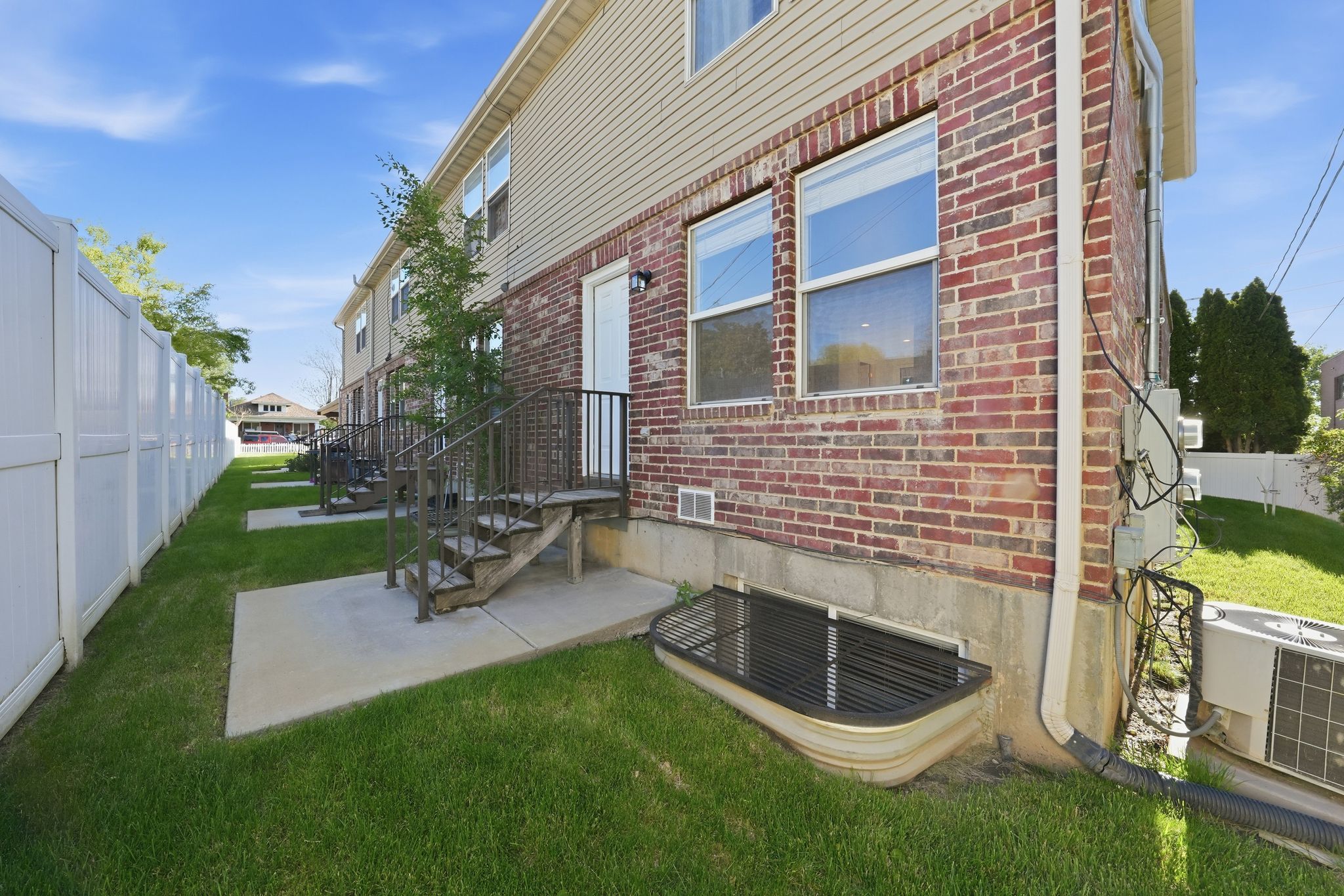 Exterior featuring a red brick facade, beige siding, white vinyl fencing, concrete patios, and a metal staircase with railings