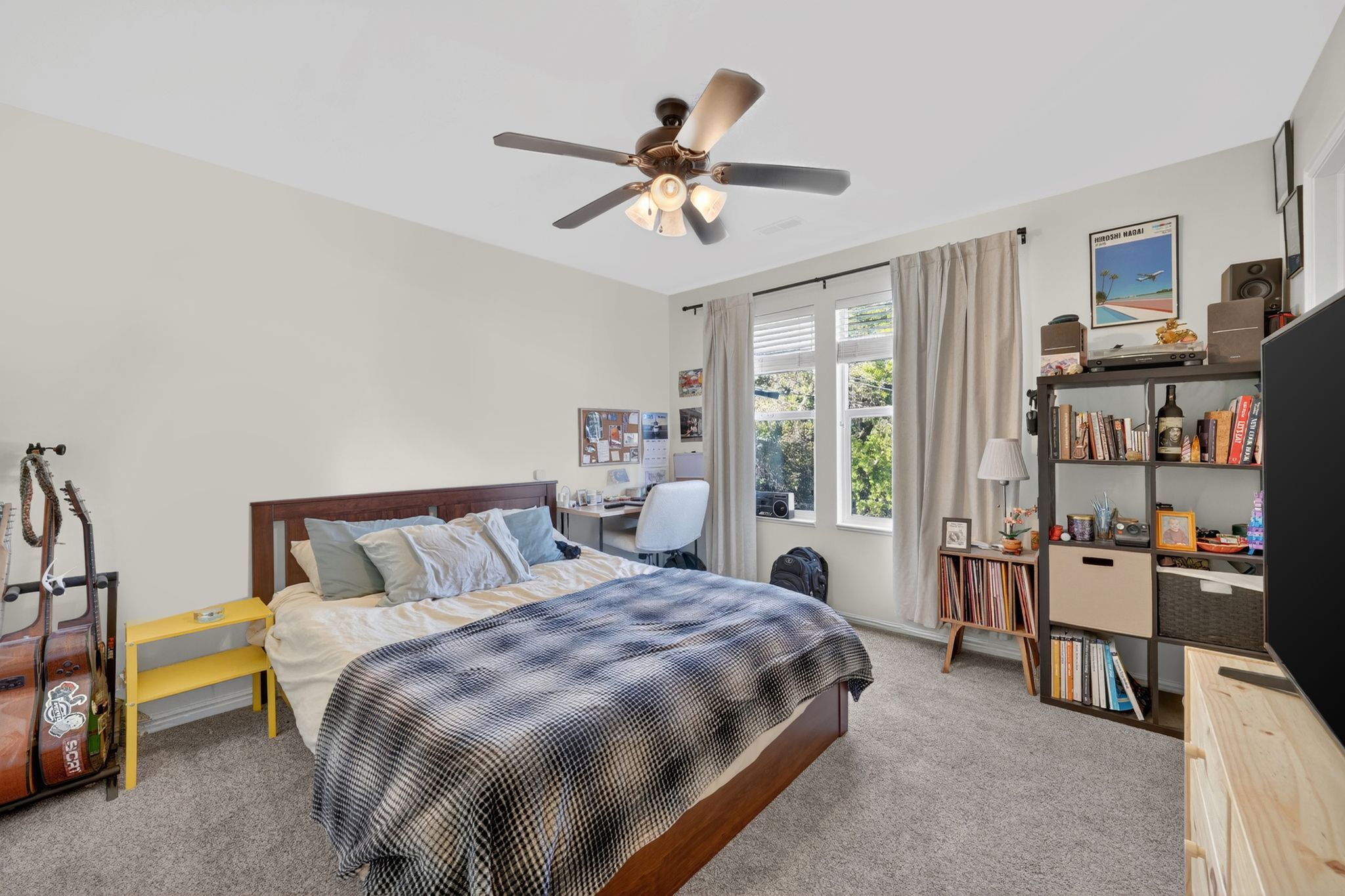 Carpeted bedroom with a ceiling fan light fixture, white walls, and a large window with white blinds and light-colored curtains