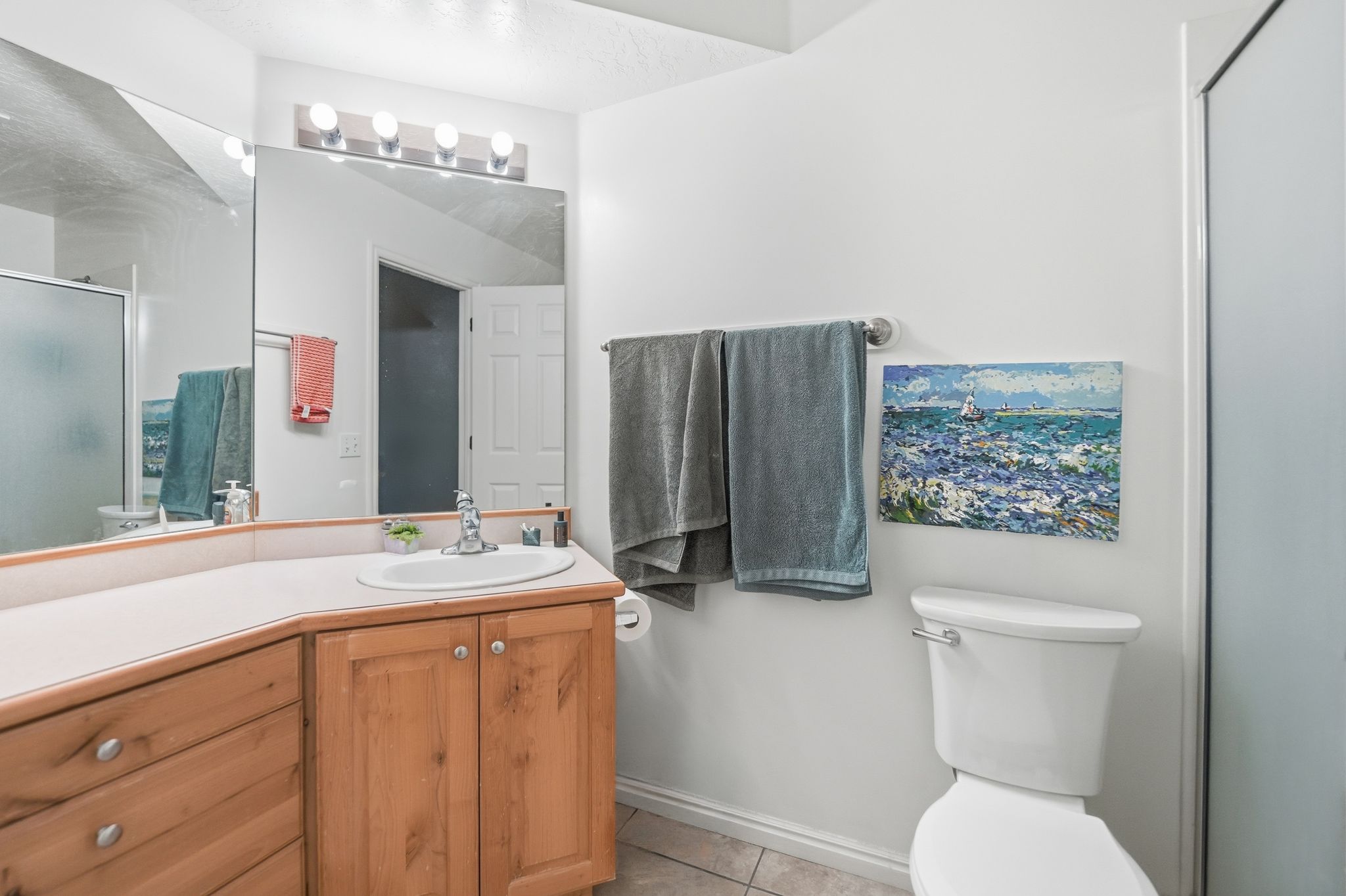 Bathroom featuring a light wood-finish vanity with a single basin, a large wall-mounted mirror, a four-light vanity fixture, a toilet, and a separate shower enclosure with a frosted glass door