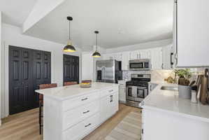 Modern kitchen featuring a large central island with a solid surface countertop, white cabinetry with dark hardware, stainless steel appliances, and wood-finish flooring