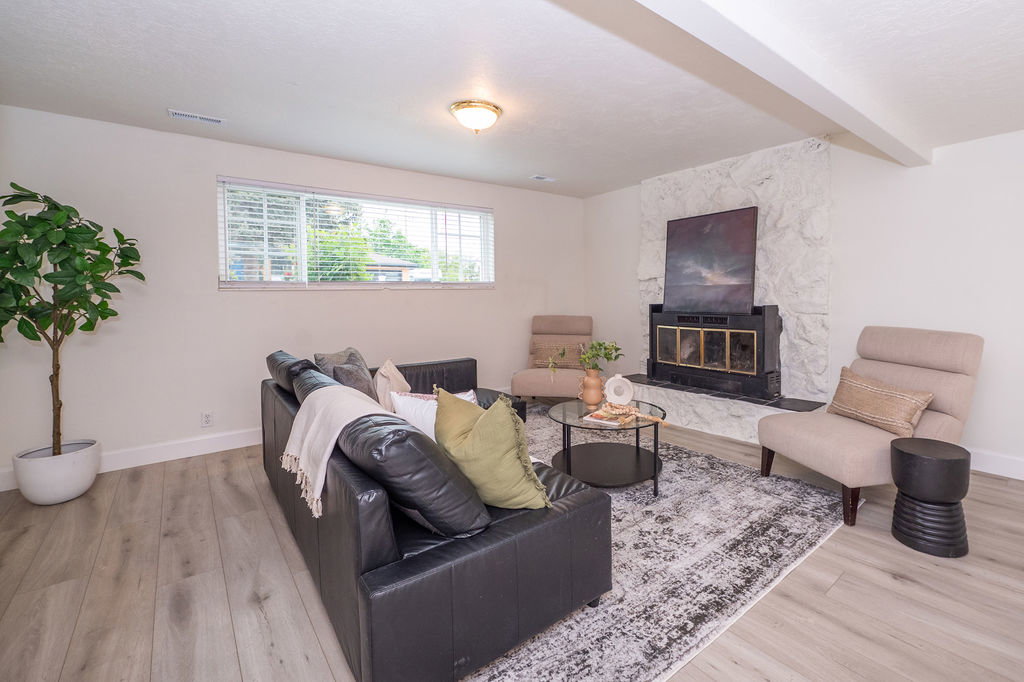 Living area featuring wood-finish flooring, a white stone-clad fireplace with a black insert, a large window with horizontal blinds, white walls, and a recessed ceiling light fixture