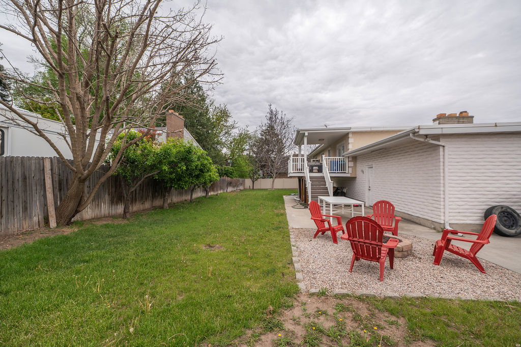 Expansive backyard featuring a gravel patio with a built-in fire pit, a mature tree line along the perimeter, a wooden privacy fence, and a concrete patio beneath an upper-level deck with white railings