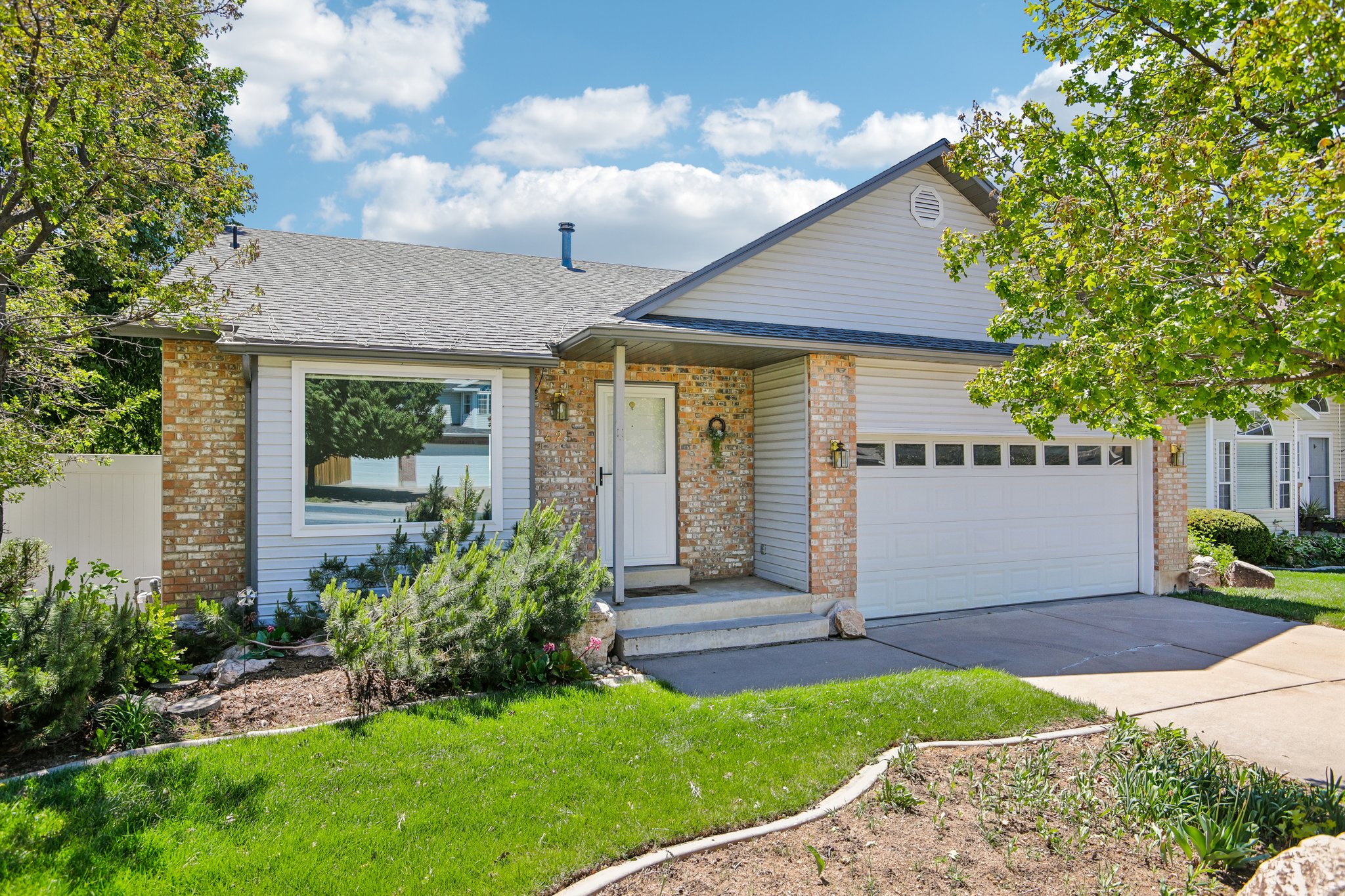 Exterior featuring brick and siding finishes, a two-car garage with transom windows, a front entry with porch lighting, and a well-maintained lawn with mature trees and landscaping