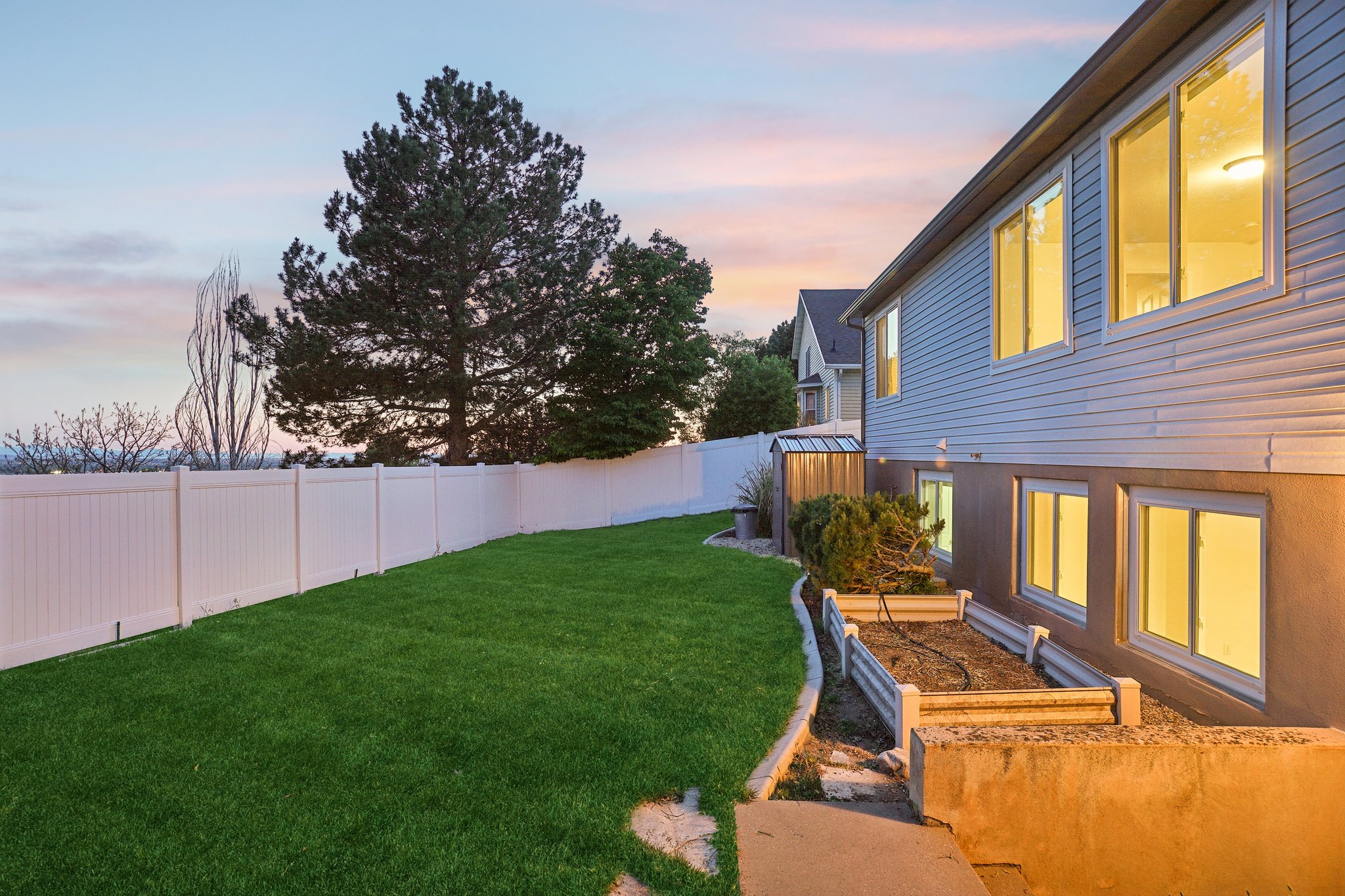 Expansive backyard featuring a manicured lawn, white vinyl privacy fencing, a dedicated garden bed, a small storage shed, and a stone-edged planting area
