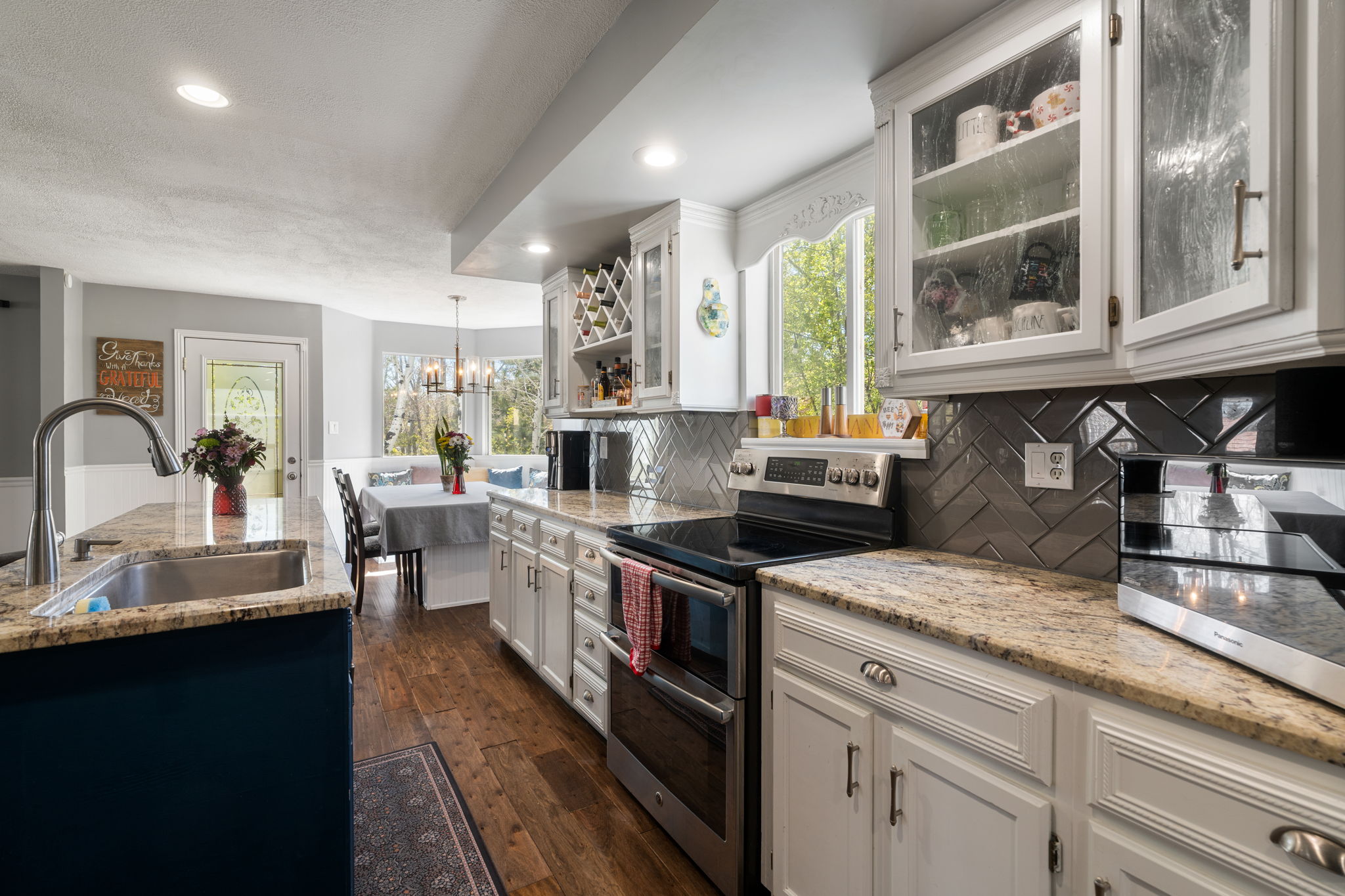 Kitchen featuring a stainless steel sink, granite countertops, white cabinetry with glass-front uppers, a herringbone tile backsplash, and wood-finish flooring