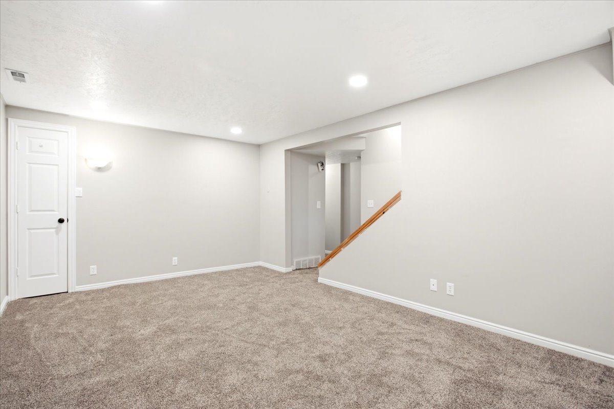 Spacious room featuring neutral carpet flooring, recessed lighting, a white panel door, and a wooden stair railing