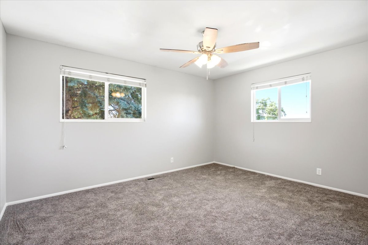 Spacious room featuring neutral carpeting, two large windows with blinds, a ceiling fan with integrated lighting, and light gray wall tones
