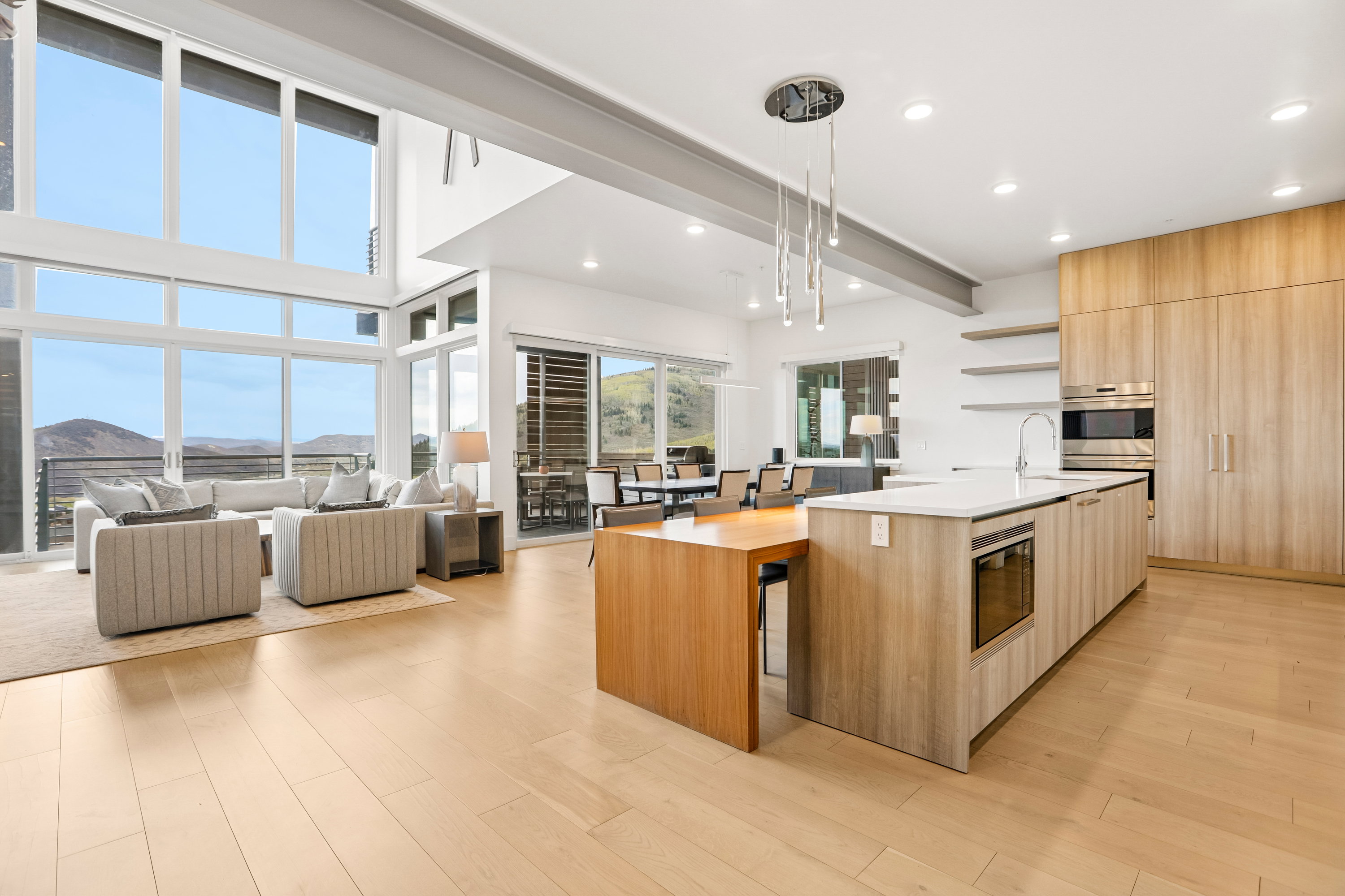 Modern open-concept living area featuring a kitchen island with integrated dining, light wood-finish flooring, and a two-story window wall