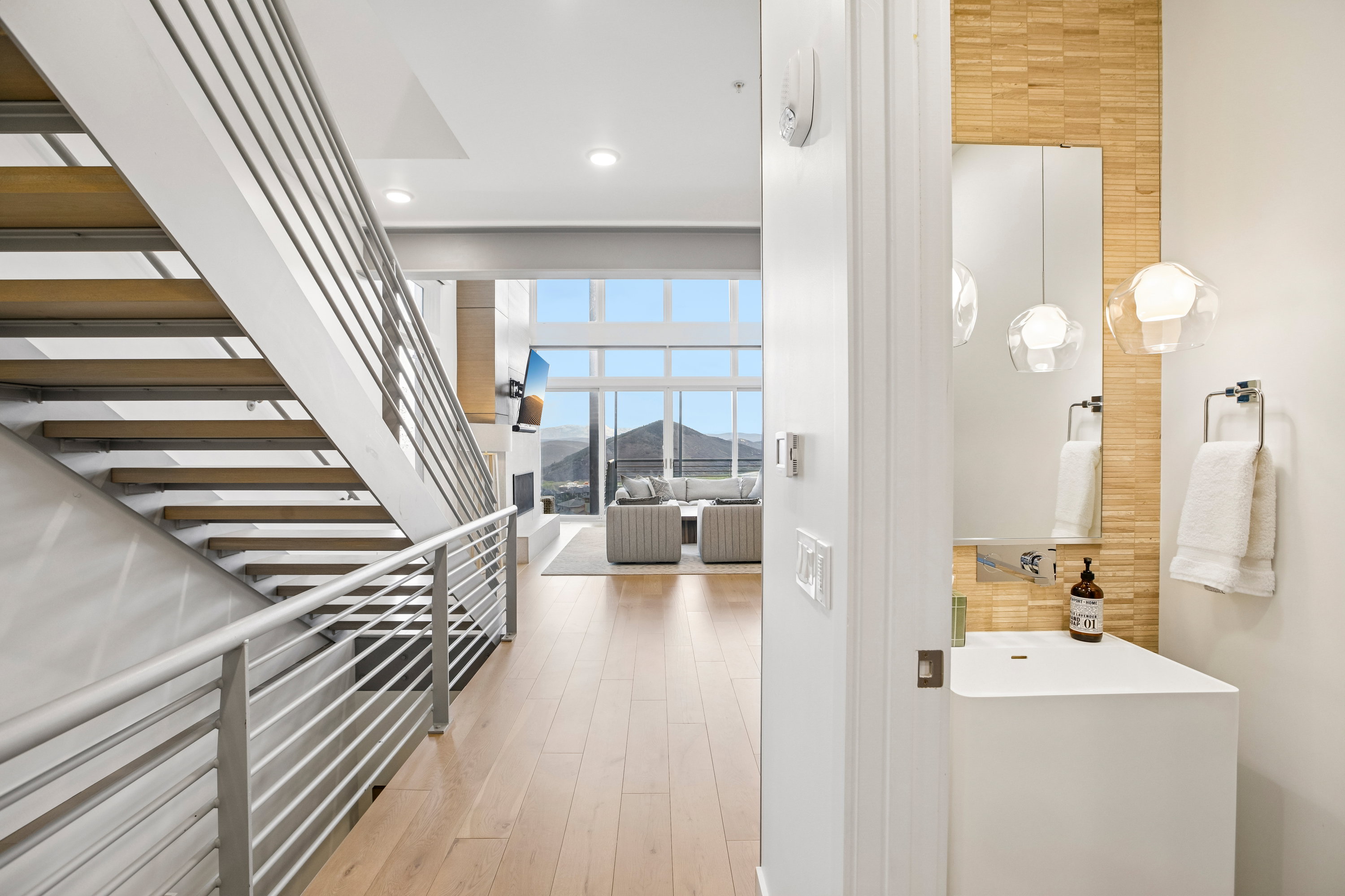 Modern staircase with horizontal metal railings, wood-finish flooring, a contemporary powder room featuring a floating vanity with integrated sink, and an expansive living area with floor-to-ceiling windows