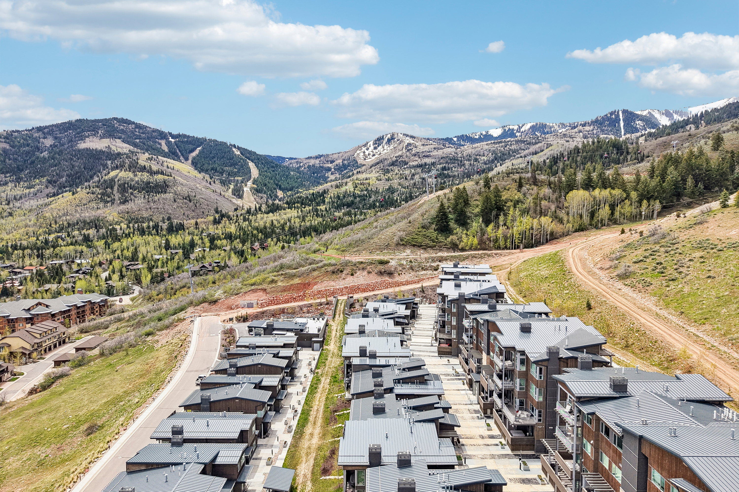 Contemporary multi-story buildings featuring dark paneling and balconies, situated amidst rolling hills with visible ski slopes and snow-capped peaks