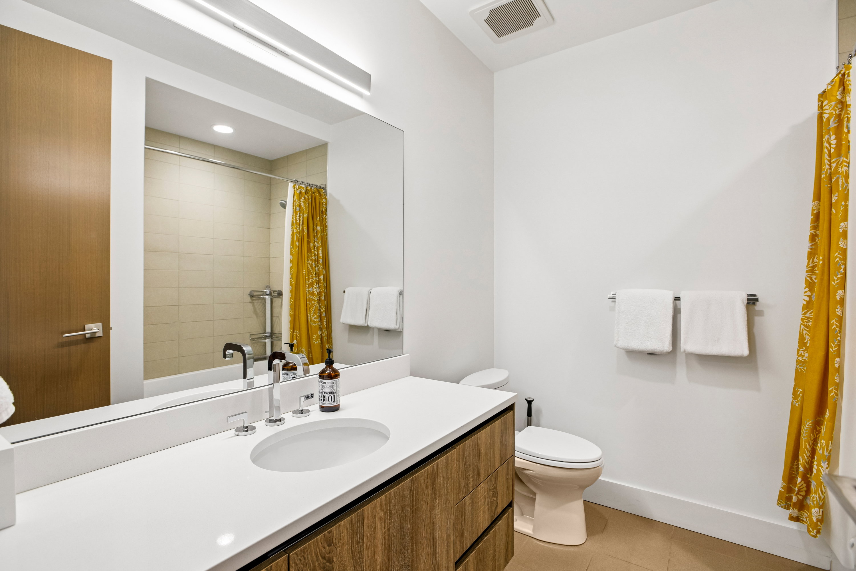 Bathroom featuring a wood-finish vanity with a white countertop and integrated sink, a large wall-mounted mirror with overhead lighting, and a bathtub with tiled surround