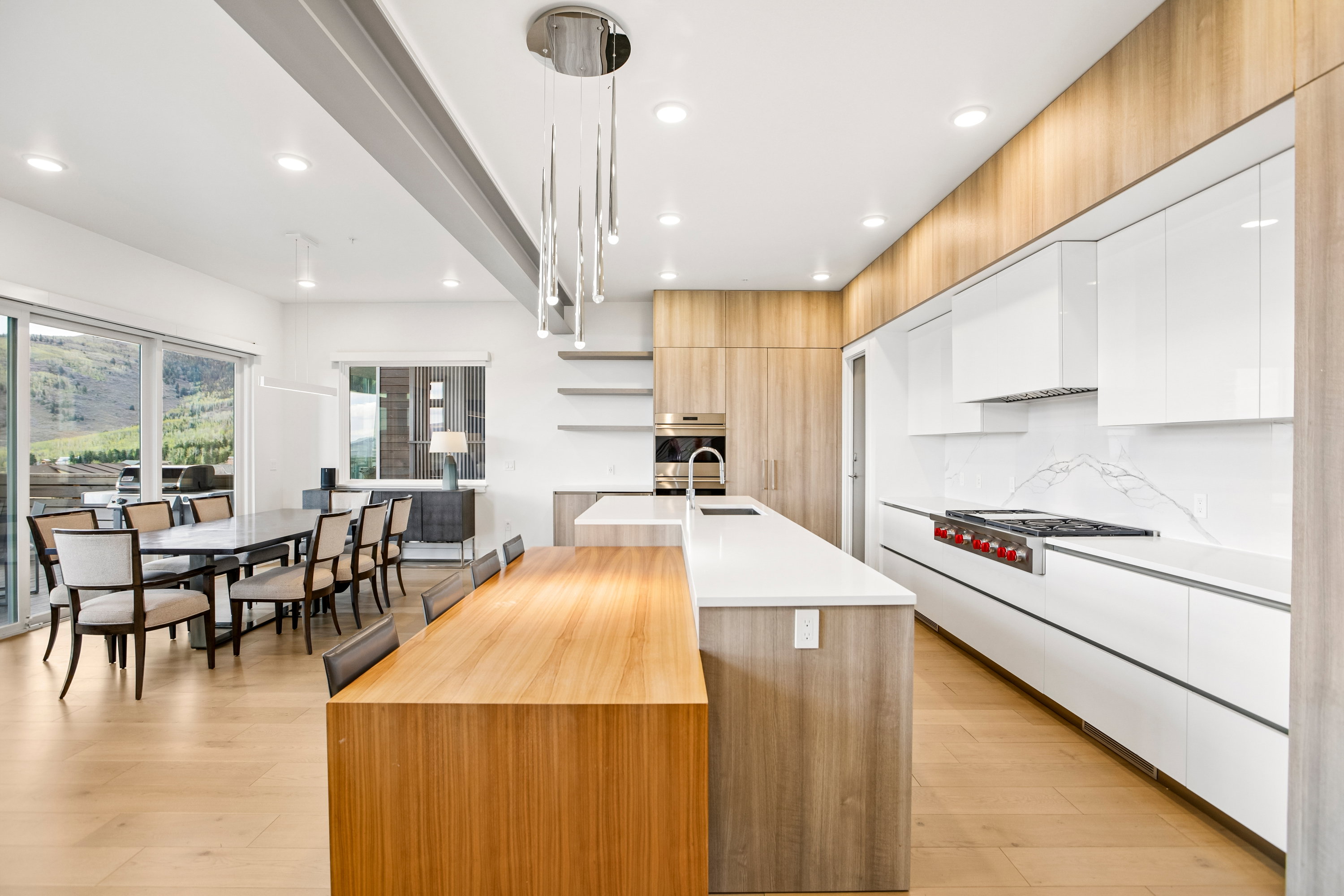Gourmet kitchen and dining area featuring a lengthy island with wood-finish paneling, light wood-finish flooring, white flat-panel cabinetry, a stainless steel range, and a full-height marble backsplash