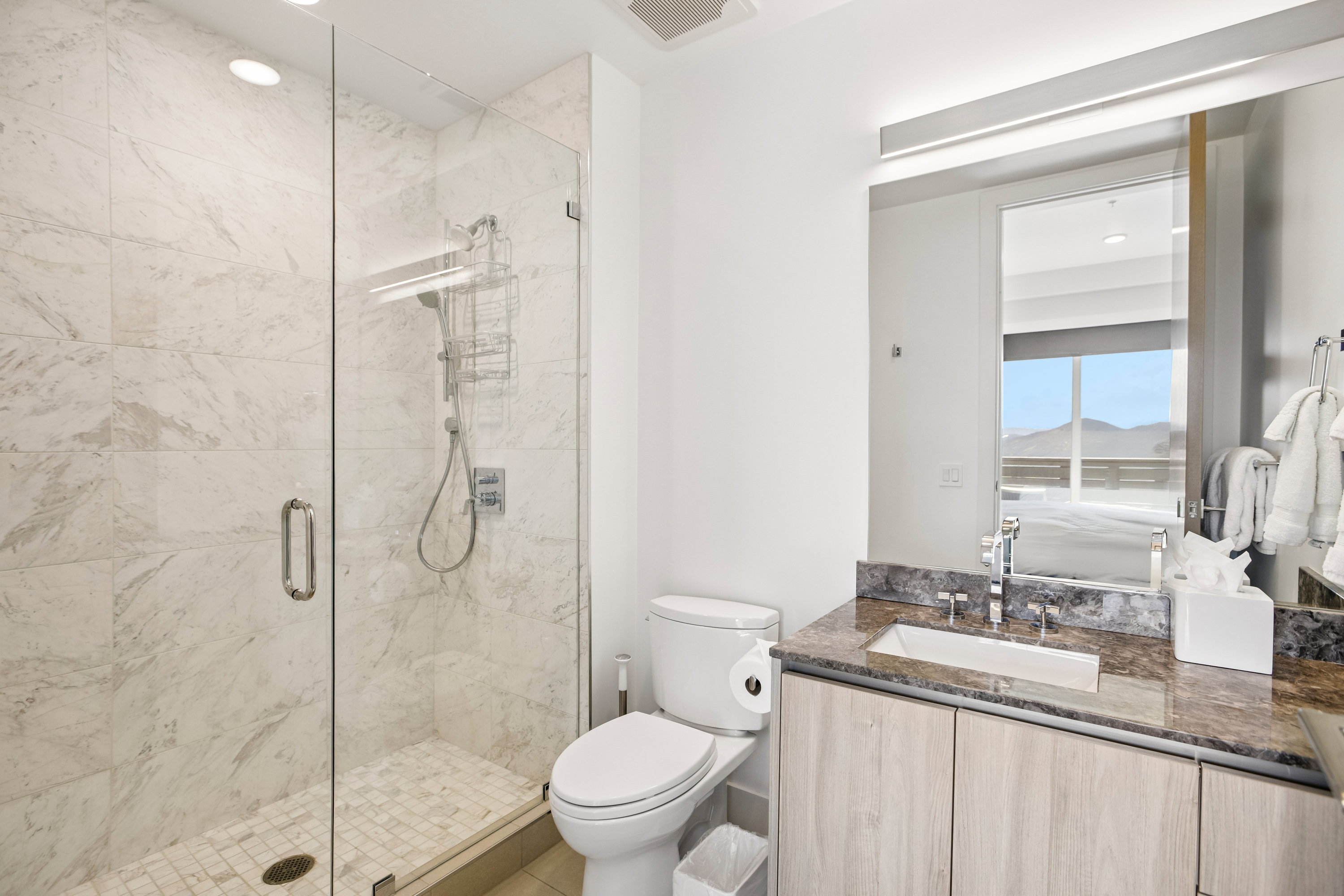 Contemporary bathroom featuring a glass-enclosed shower with large format wall tiles and a mosaic tile floor, an integrated vanity with wood-finish cabinetry, a stone countertop, and a rectangular undermount sink with a wall-mounted mirror