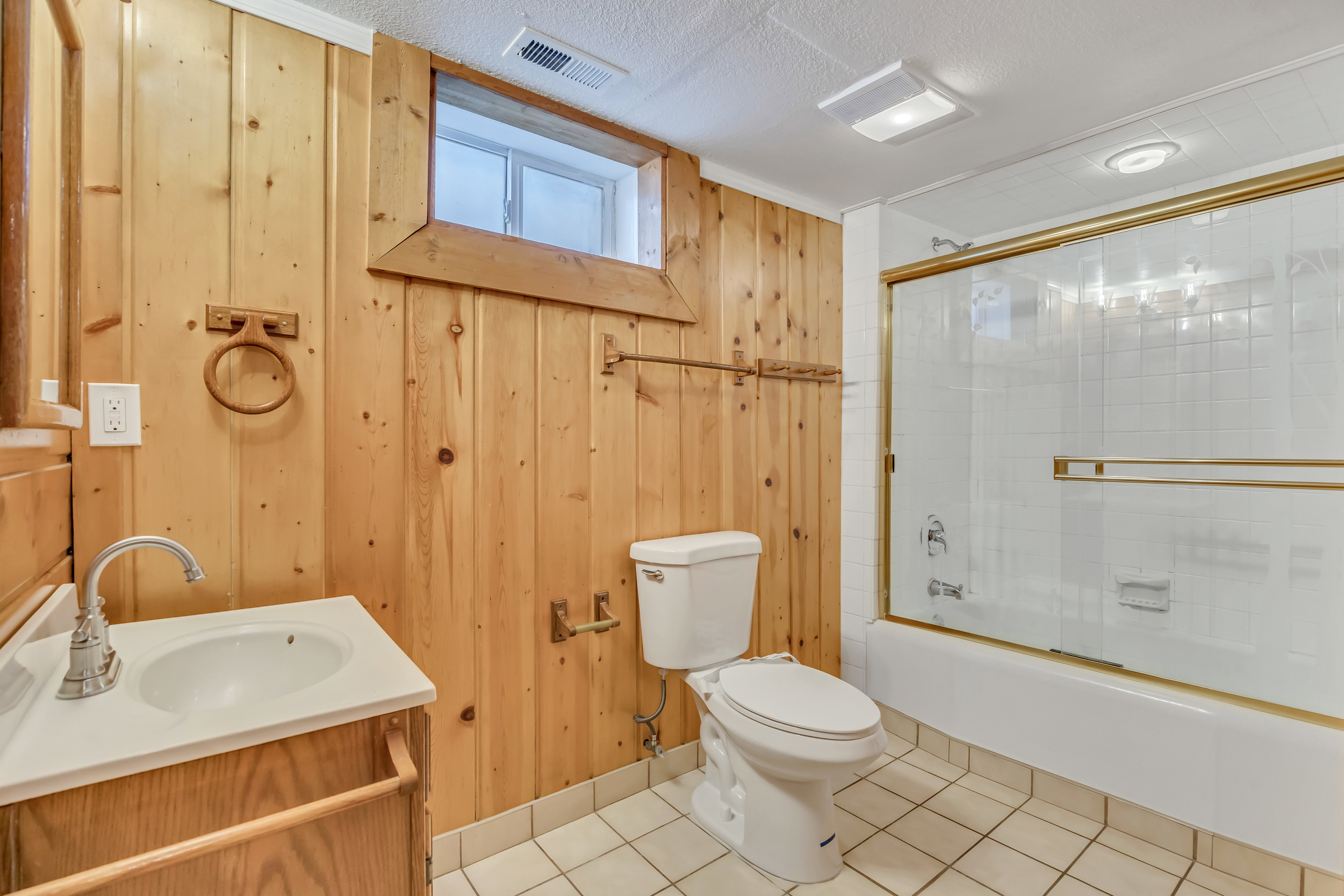Bathroom featuring wood paneling, a white vanity with an integrated sink, a toilet, and a bathtub with a gold-framed sliding glass door