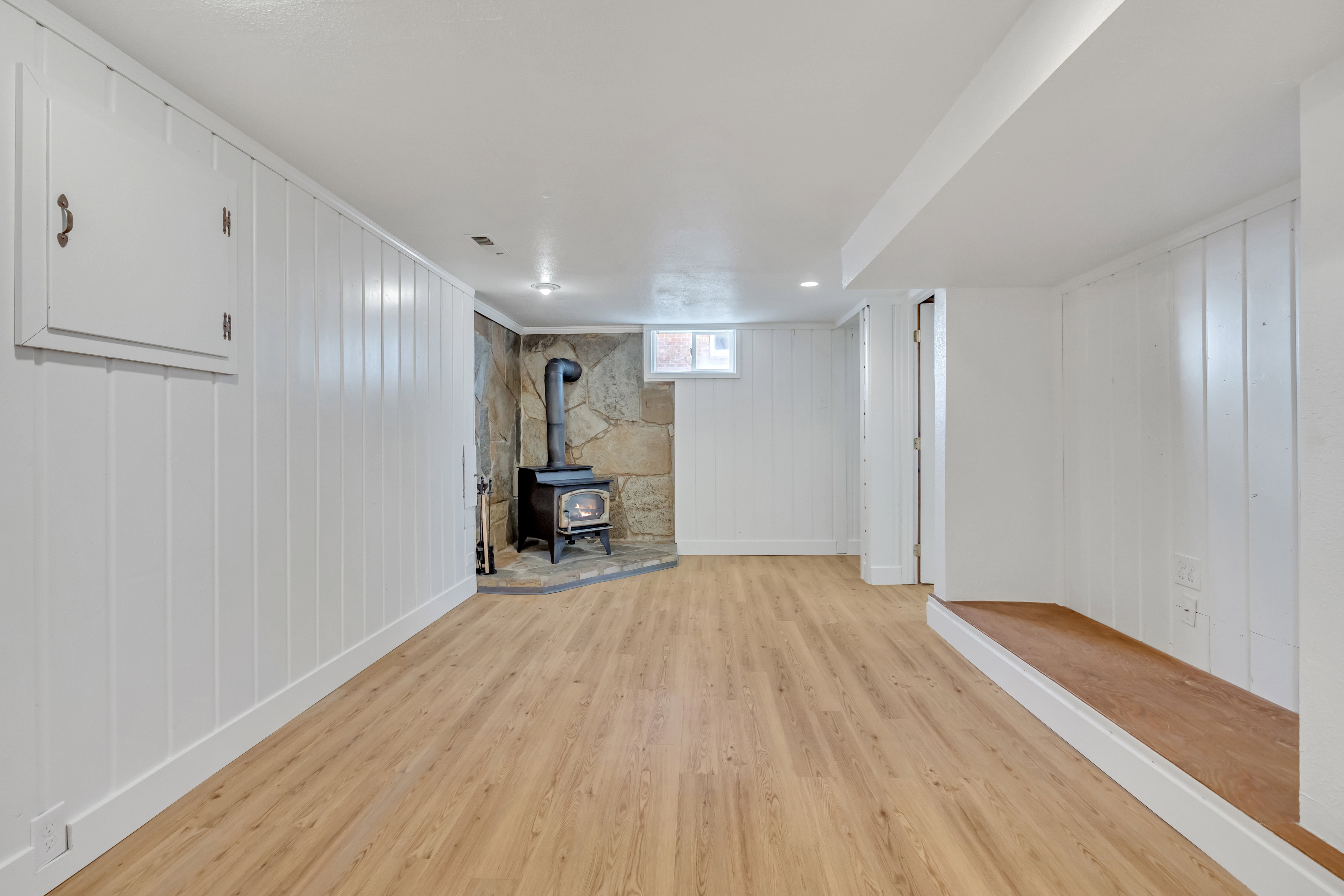 Finished basement area featuring a wood-burning stove with a stone surround, wood-finish flooring, white paneled walls, and recessed lighting