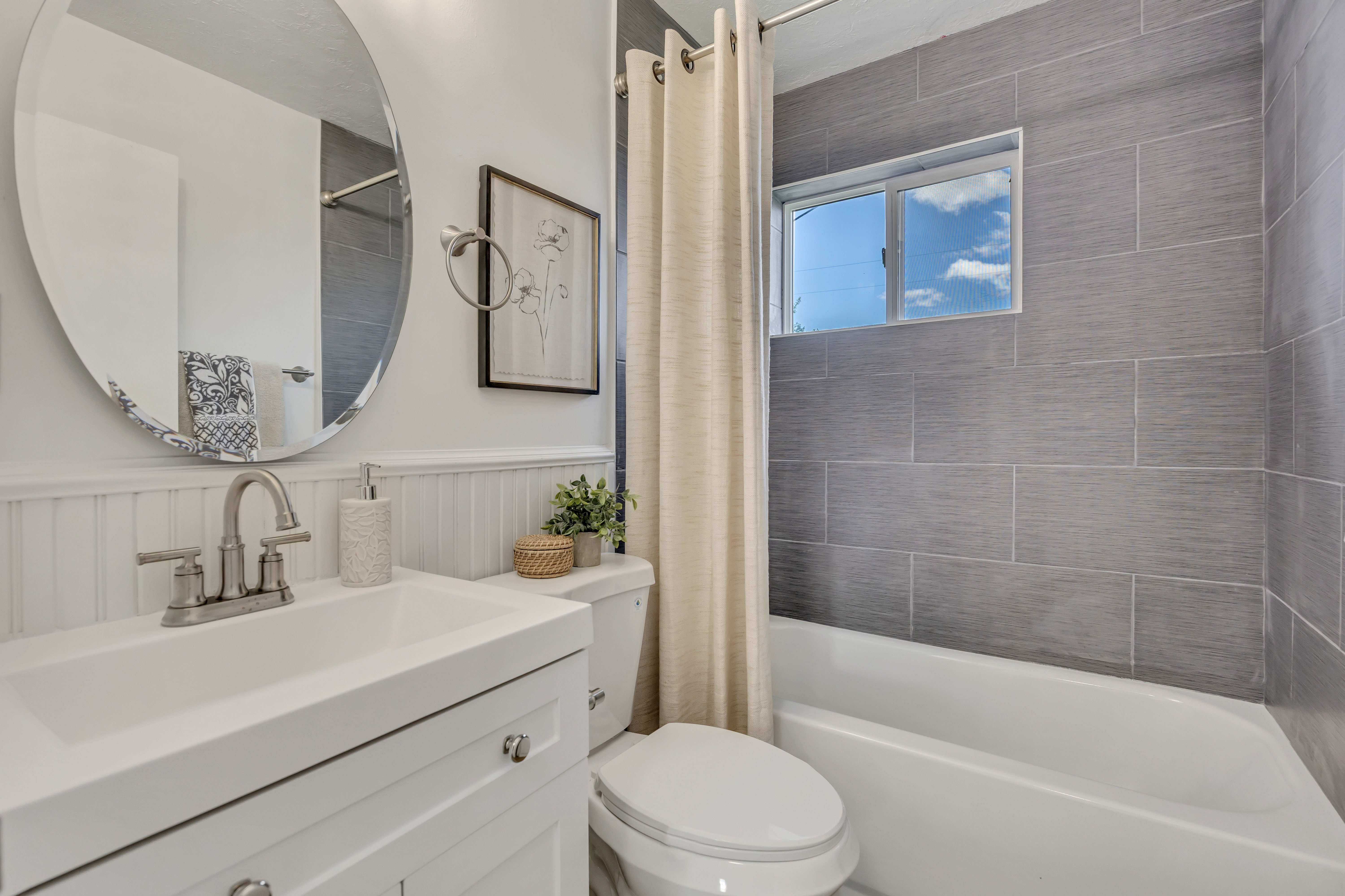 Bathroom featuring a white vanity with integrated sink, brushed nickel faucet, beadboard wainscoting, and a tiled shower/tub combination with a window