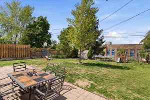 Expansive backyard featuring a paver patio with a checkerboard-patterned tabletop, a lush lawn, mature trees, a wooden privacy fence, and a white storage shed