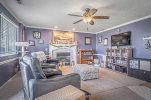 Living area with a ceiling fan light fixture, crown molding, and a fireplace featuring a mantel
