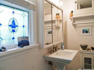 Stained-glass window with a fleur-de-lis motif, white pedestal sink, dual-handle faucet, mirrored medicine cabinet, and wall-mounted shelving