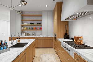 Gourmet kitchen featuring a large island with an undermount sink, marble countertops, natural wood-finish cabinetry, a professional-grade range with a custom hood, and open shelving