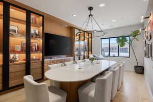 Spacious room featuring a large oval table with a light-colored surface, built-in display cabinetry with glass doors, integrated lighting, a large wall-mounted screen, and light wood-finish flooring