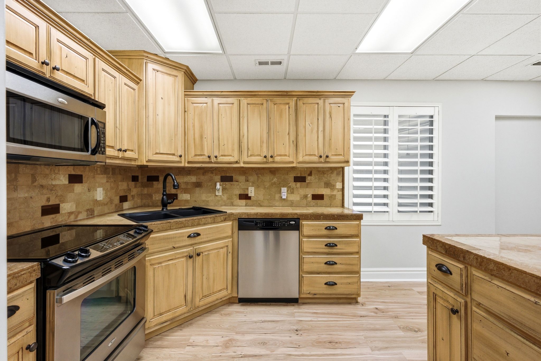 Kitchen with stainless steel appliances, tile countertops, light wood-style floors, a drop ceiling, and light wood finish cabinetry