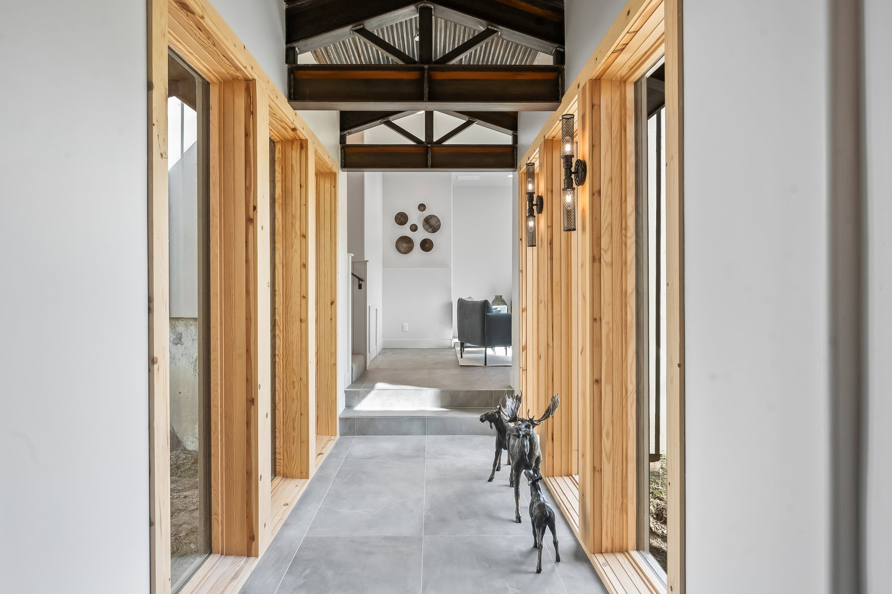 Hallway featuring wood-framed interior windows, exposed structural beams, corrugated metal ceiling, contemporary wall sconces, and large format tile flooring