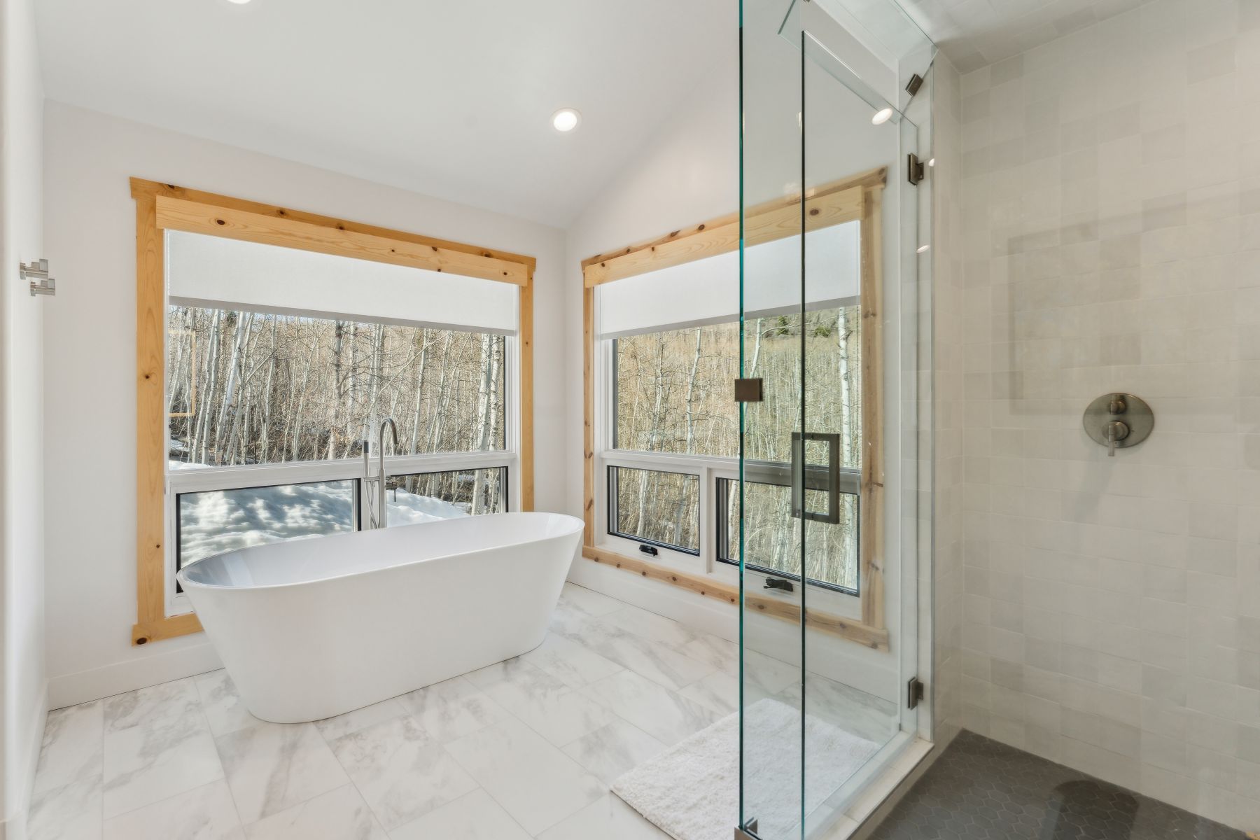 Freestanding white soaking tub with a floor-mounted faucet, set against large windows with natural wood trim