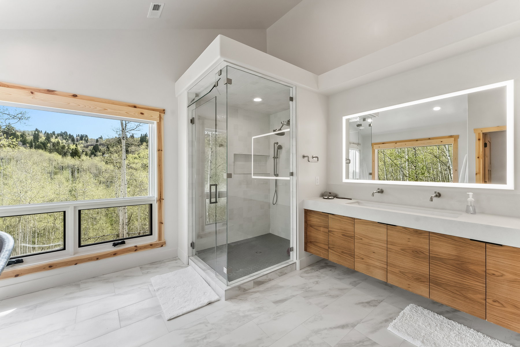 Contemporary bathroom featuring a spacious glass-enclosed shower, a double vanity with a light wood-finish base and dual vessel sinks, and an illuminated mirror