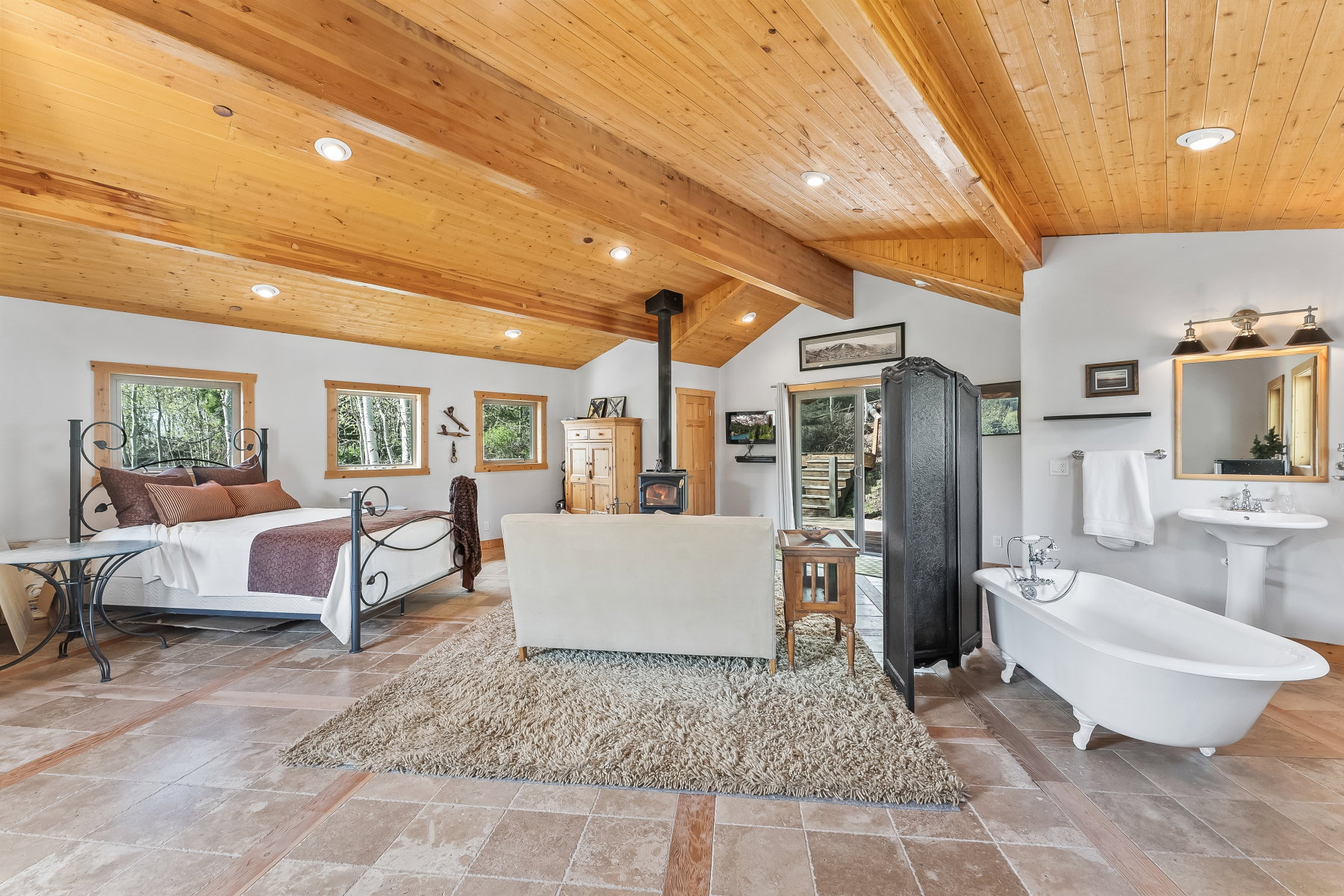 Vaulted wood-plank ceilings with exposed beams, wood-frame windows, a freestanding clawfoot tub, and a wood-burning stove