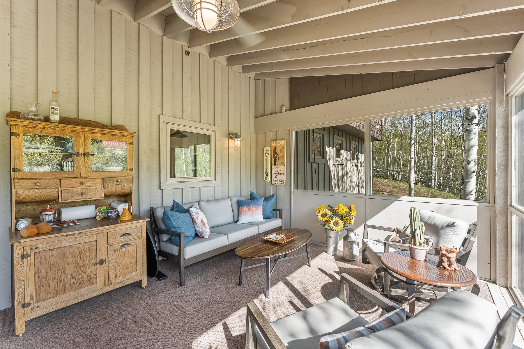 Screened porch featuring a natural wood built-in hutch, exposed beam ceiling, wall-mounted sconce lighting, and a tree-lined backdrop