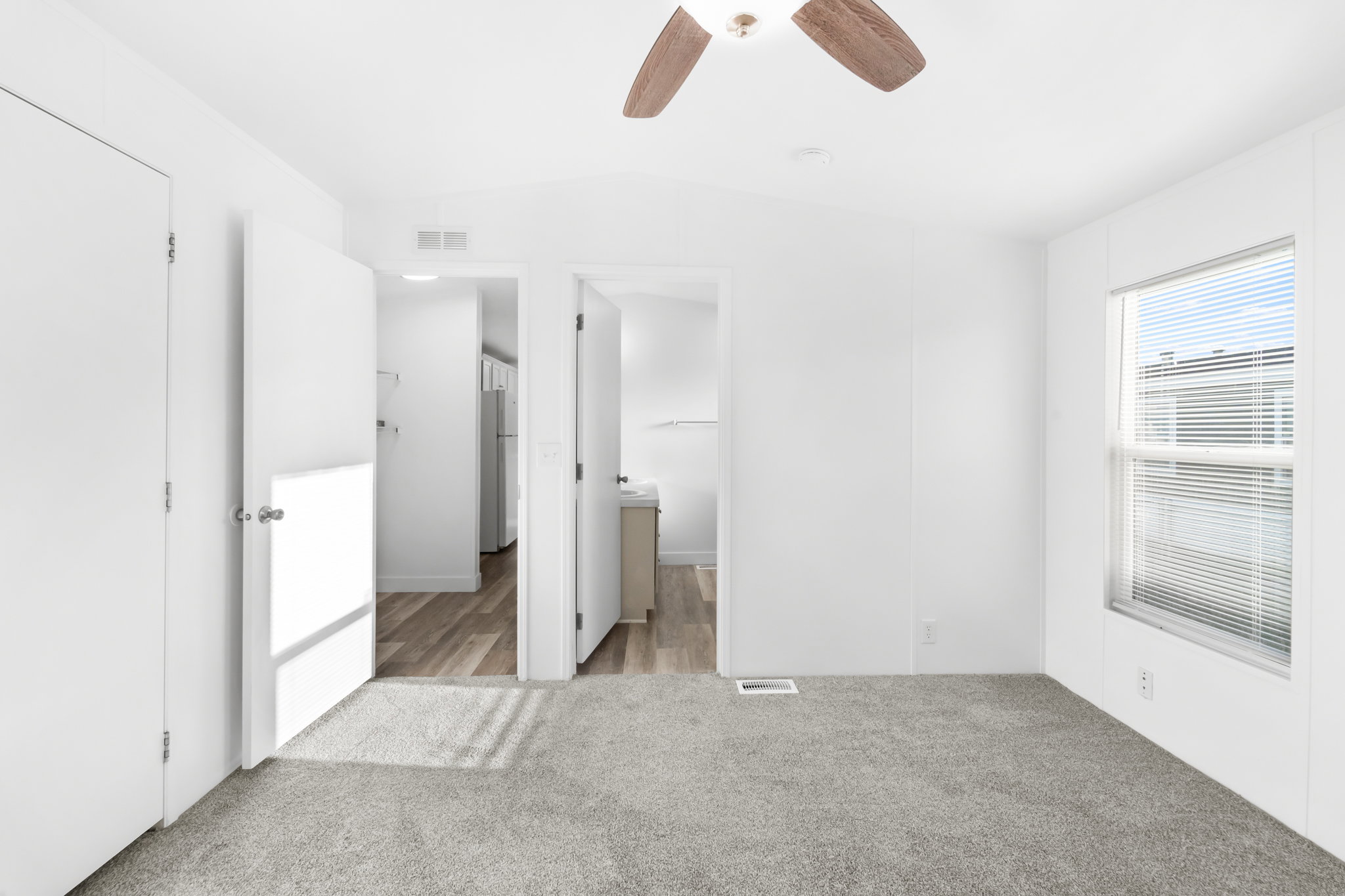 Neutral white interior featuring light gray carpeting, a window with horizontal blinds, and a ceiling fan with wood-finish blades