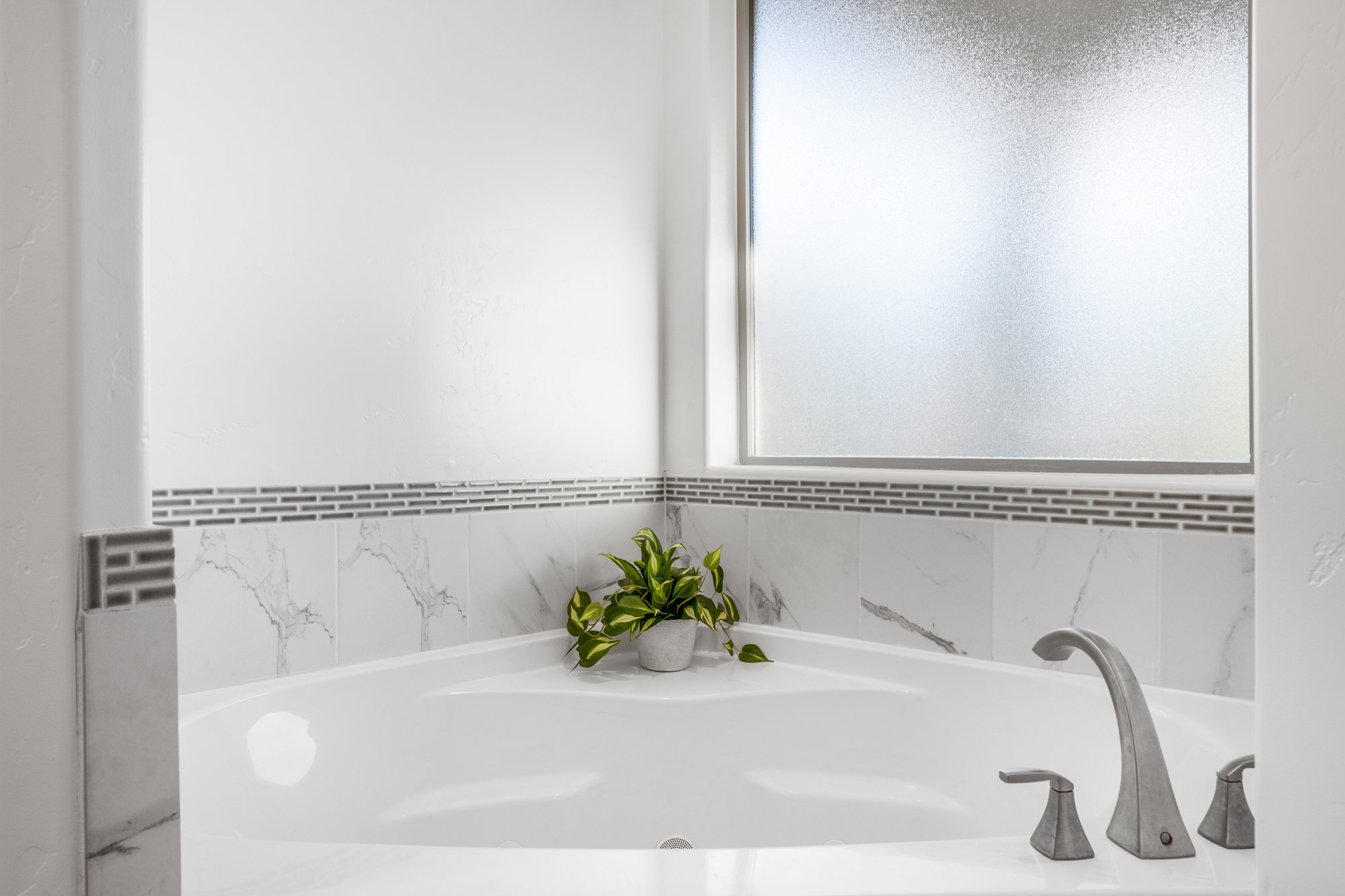 Built-in soaking tub featuring a matte nickel faucet, marble-look tile surround, decorative mosaic tile accent, and a privacy window