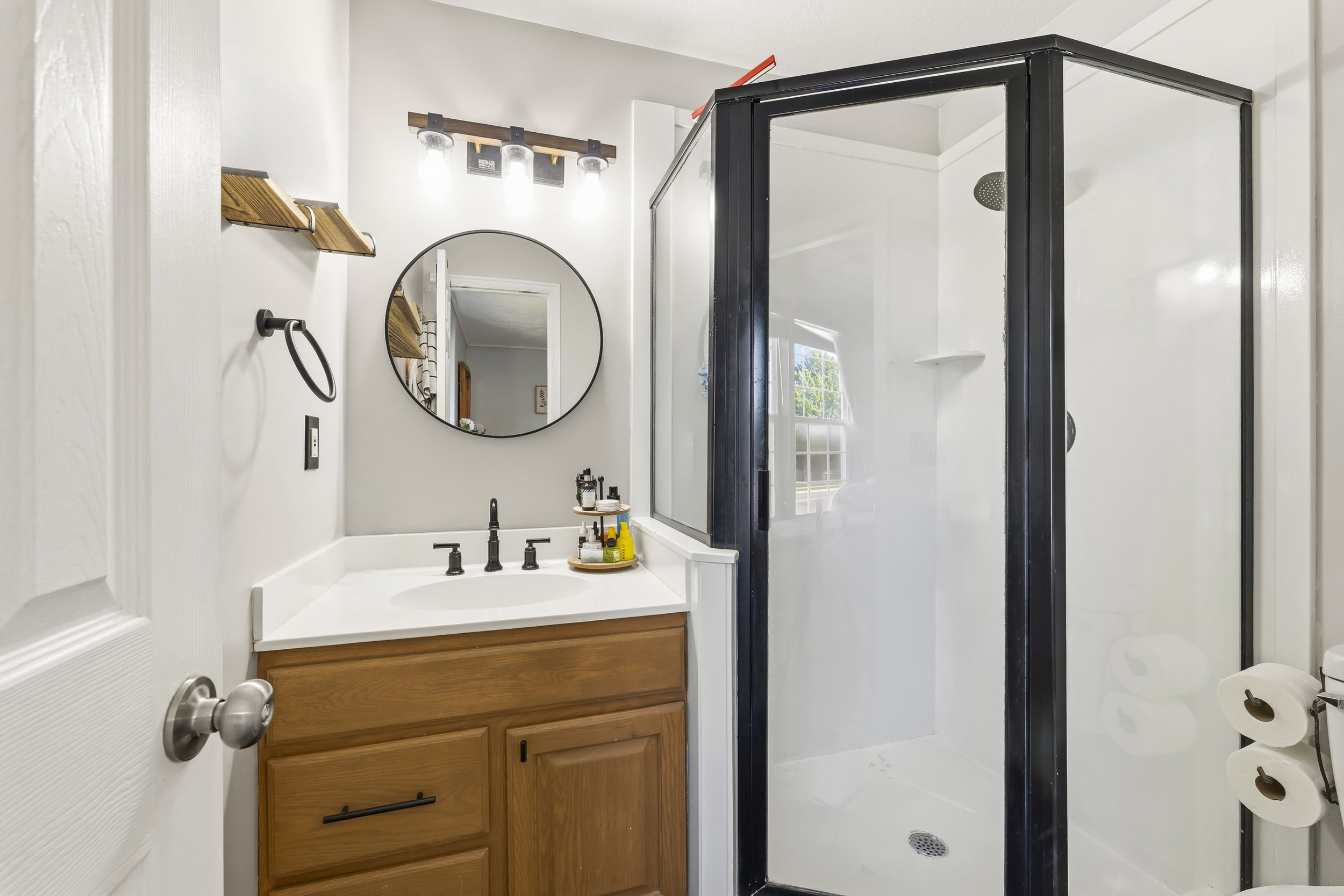 Full bathroom featuring a walk-in shower with black framing, a single vanity with wood-finish cabinetry, a white countertop, and a circular mirror