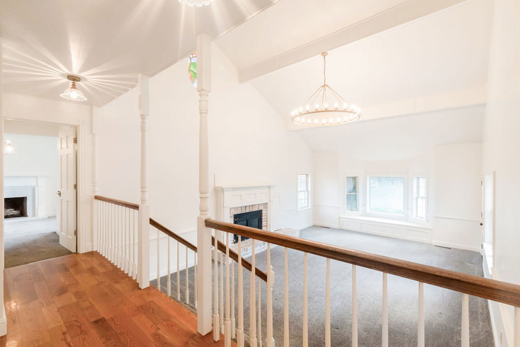 Vaulted ceiling great room featuring a grand chandelier, bay window with integrated seating, and a fireplace with a white mantel, visible from the upper-level hallway with wood-finish flooring and white railings