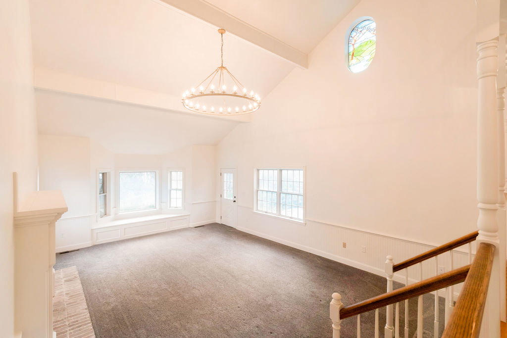 Spacious living area featuring a vaulted ceiling with exposed beams, a large circular chandelier, and a decorative stained glass oval window