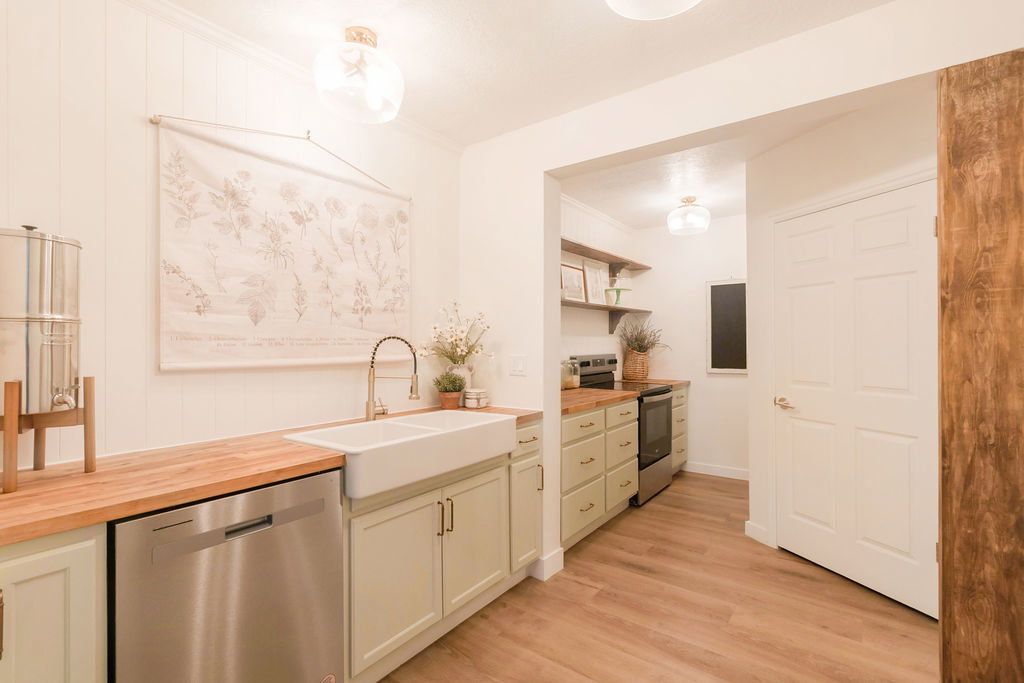 Kitchen featuring wood-finish flooring, butcher block countertops, light green cabinetry, a white farmhouse sink with a gooseneck faucet, and a stainless steel dishwasher