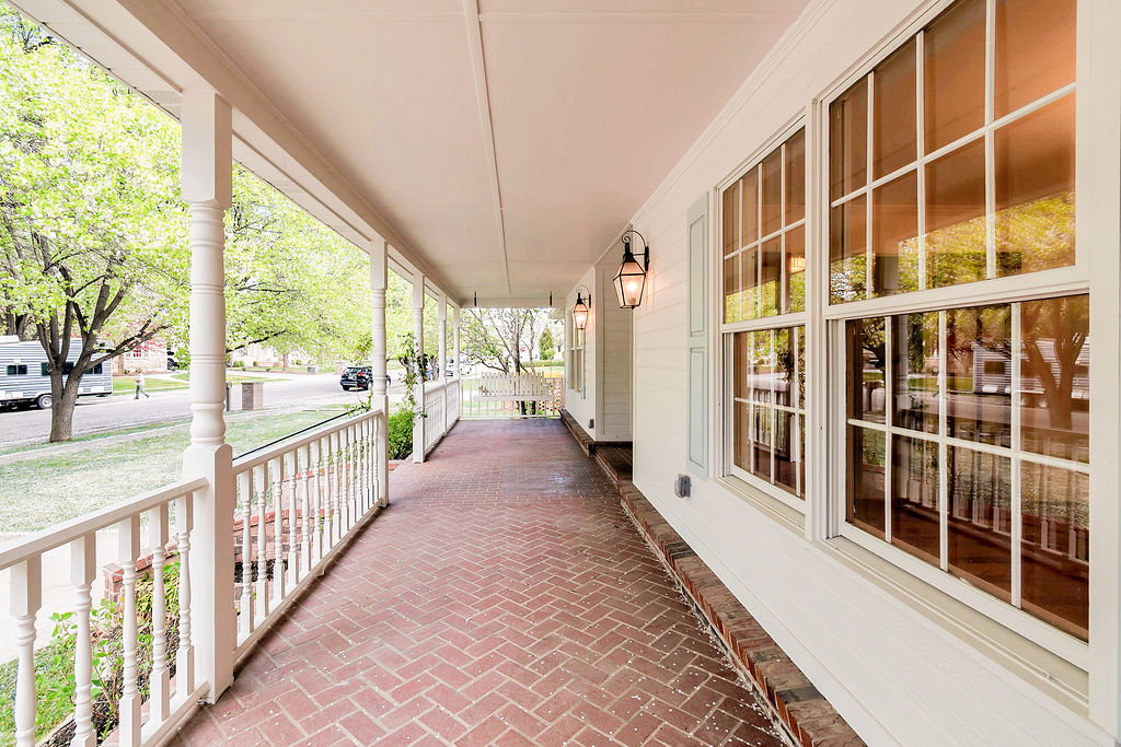 Expansive covered front porch featuring a herringbone brick pattern floor, white railing, decorative columns, and exterior wall lanterns